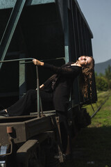 a woman in a black suit posing against the background of a railway carriage. She stands on the metal platform of the carriage, holding the handrail with one hand.