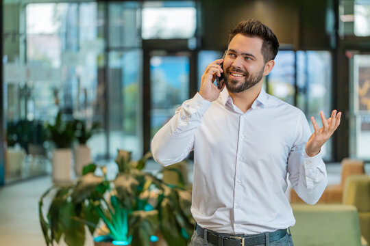 Smiling happy businessman in formal shirt working, having mobile phone talk in modern office lobby. Professional manager man guy holding smartphone using messenger chat apps. Employment, occupation