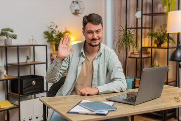 Hello. Happy Caucasian man freelancer waves hands hi gesture greeting welcomes someone, online webinar, remote distance conversation. Freelance guy sitting at table desk with laptop in home office.