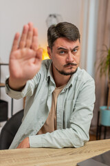 Young man freelancer with serious expression doing stop gesture with hand at home office desk,...