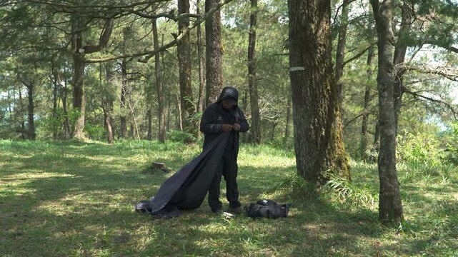 A solo traveler is preparing a flysheet tent in a green and lush pine forest area. Asian man adventuring alone in the jungle and setting up a trap tent from flysheet for bushcraft and camping
