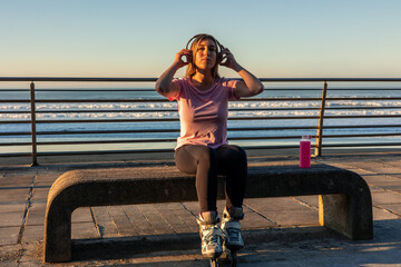Young woman with inline skates listening music on a bench by the sea at sunset