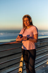 vertical Sportswoman using smartphone and listening music with headphones at sunset on the beach