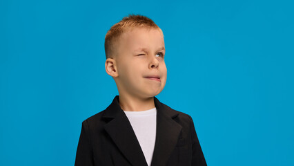 Playful boy in black blazer and white shirt, winking with lips pursed slightly, showcasing a cheeky, fun attitude against bright blue background. Concept of childhood, beauty, fashion, style. Ad © Lustre Art Group 