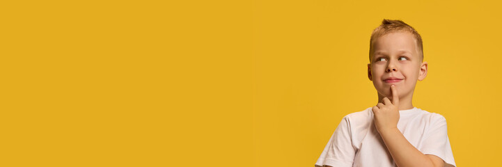 Boy strikes thoughtful pose, hand on chin, smiling slyly, dressed in white shirt, exuding curiosity against bright yellow background. Concept of childhood, beauty, fashion, style. Ad