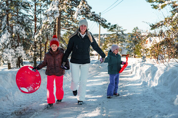 Smiling mother and two daughters walking down a snowy road on a bright winter day, joyfully holding snow discs and creating cherished memories together in the winter wonderland