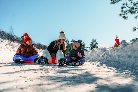 Mother and daughters sitting on snow sleds, taking a joyful break from sledding while basking in the warmth of a sunny winter day amidst the majestic mountains