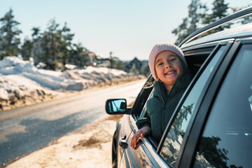 Happy child leaning out of the car window, grinning widely while enjoying a winter road trip. Snow-covered landscapes create a beautiful backdrop for this joyful adventure