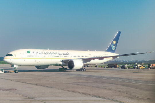 Algiers, Algeria- December 29, 2024: Saudi Arabian company airplane Boeing 777, trucks and utility vehicles parked on the airport tarmac outdoors, sunlights shining in a sunny day with clear blue sky.