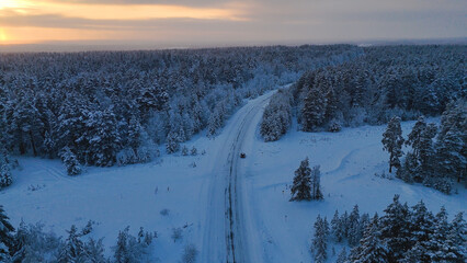 The car is driving along the road in a snow-covered white spruce forest at sunset