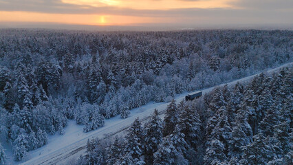 The car is driving along the road in a snow-covered white spruce forest at sunset