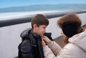 A woman helps her son by buttoning his coat while standing outside near the seaside. The scene...