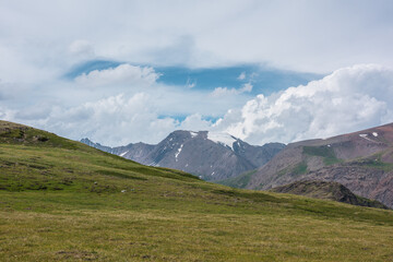 Dramatic alpine view from green hills to large rocky mountain range with glacier under clouds in blue sky. Scenic landscape with big snow mountain top in center. Changeable weather in high mountains.