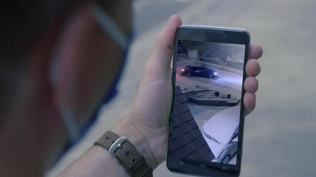 A man, perhaps while on vacation, watches security camera footage on his cellular phone as a car slides down the street outside his home.	