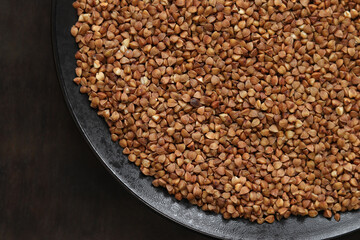 Raw buckwheat in a black plate on a dark wooden table. Buckwheat groats in a plate, top view. Raw buckwheat grains, uncooked porridge. Buckwheat background