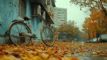 Fototapeta premium An old bicycle rests against a weathered blue wall in an autumnal city street, surrounded by fallen leaves. Foggy, muted colors and a quiet atmosphere.