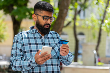 Shocked Indian man trying to pay online shopping with smartphone blocked credit card. Annoyed bearded Hispanic tourist on street buying bank refuse problem unsuccessful payment lack of money balance