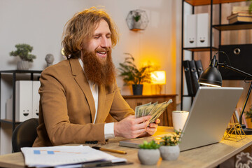 Happy Caucasian redhead businessman counting money cash and using laptop in home office. Successful...