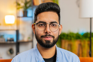 Portrait of happy Hispanic man sitting on sofa looking at camera and smiling at home. Young adult Indian bearded guy wearing eyeglasses having attractive appearance relaxing thinking in living room.