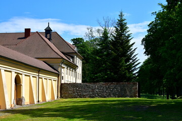 A view of an old wall, entrance gate, and tower of an old monastery, church, or shrine located next to a dense and lush forest or moor and a well maintained lawn seen on a sunny summer day in Poland