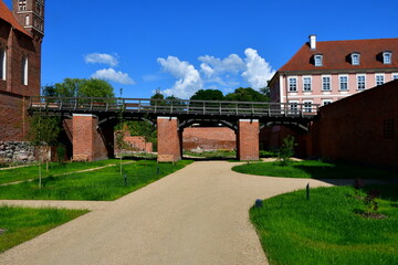 A view of a small public garden with plots of land, decorations, lighting, and paths for walking, located next to a medieval red brick wall or gate and a shallow yet vast river or lake seen in Poland