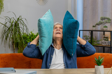 Repair work at neighbors. Irritated senior old grandfather man cover ears with pillows annoyed by noisy loud party music neighbors wishes silence. Thin walls at home flat without sound insulation