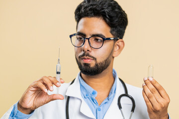 Indian young doctor cardiologist man holds syringe needle and ampoule tube with medical vaccine medicine treatment injection ready to use. Immunization. Arab scientist guy isolated on beige background