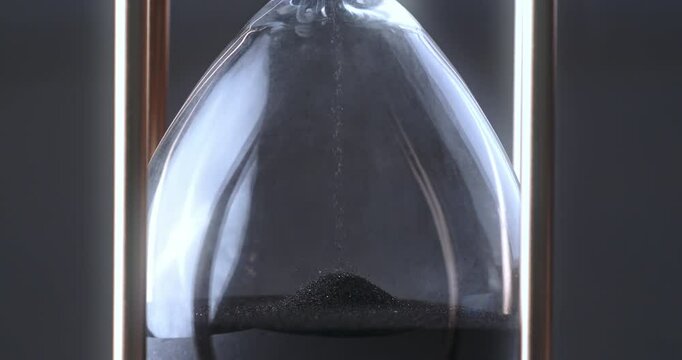 Super slow motion macro of fluid flow of black sand grains particles running inside hourglass sandglass clepsydra measuring passing time in countdown to deadline at 1000 fps.