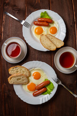 Breakfast two plates fried eggs with sausages white bread and tea in cups on a wooden table