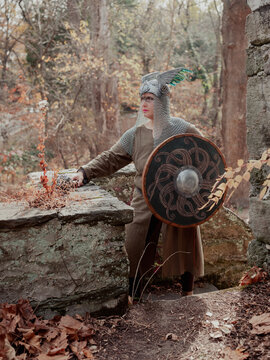 Female Viking in winged and chainmail armor and face paint standing in thoughtful pose in stone ruins. 
