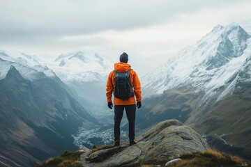 Fototapeta premium A hiker in an orange jacket stands atop a mountain, admiring the stunning view of snow-capped peaks.
