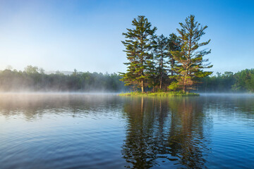 Obraz premium Beautiful calm blue lake and island with pine trees and fog on a summer morning in northern Minnesota