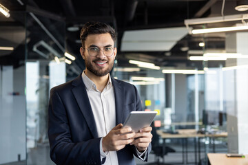 Latin American businessman dressed in formal attire holding a tablet and smiling confidently in a...