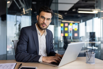 Confident Latin American businessman using a laptop while seated in a contemporary office environment. Focused professional showcasing productivity and determination in a workspace.
