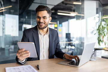 Latin American businessman working on tasks using a tablet and laptop in a modern workspace,...