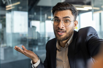 Obraz premium Latin American businessman wearing glasses and suit engaged in animated conversation in a contemporary office space, expressing opinions confidently with hand gestures.