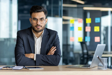 Latin American businessman wearing glasses in an office sitting at a desk with arms crossed expressing confidence. Visible items include a laptop, and notes, indicating a professional environment.