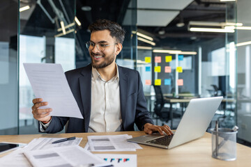 Obraz premium Latin American businessman reviews important documents while working on his laptop in a modern yet professional office environment. This portrays professionalism, productivity, and a positive work