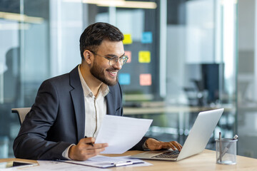 Latin American businessman in a formal suit smiling while working on a laptop in a modern office, holding documents and sitting at a desk surrounded by professional workspace items.