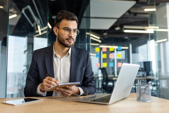 Latin American businessman attentively working at a desk with a laptop, notebook, and pen reflecting professionalism and focus in a modern office setting surrounded by contemporary work