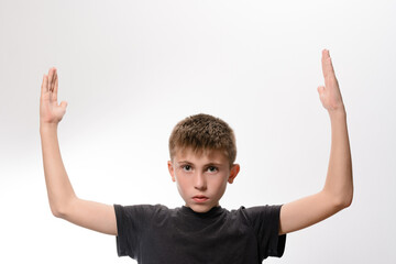 A young boy with short brown hair stands against a plain white background, his arms raised above...