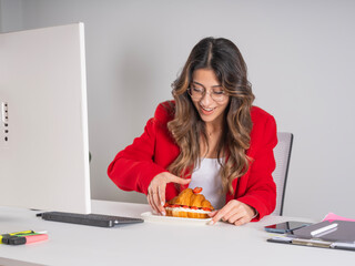 Happy hungry businesswoman,  portrait of young excited happy hungry businesswoman eating fresh delicious croissant while sitting on office desk wear red formal jacket. Breakfast concept image.