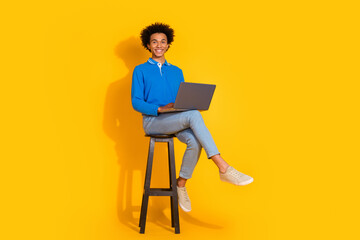 Full size photo of pretty young male sit wooden chair netbook wear trendy blue outfit isolated on yellow color background
