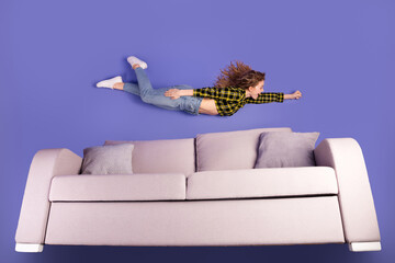 Young woman simulating flying superhero pose above a sofa against a purple background