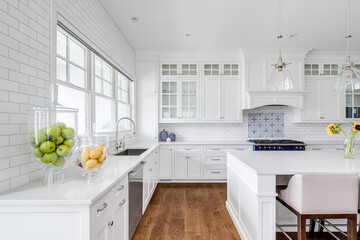 A white kitchen detail with bar stools sitting at a large island, glass lights hanging from the ceiling, and a beautiful tiled backsplash. No brands or labels.