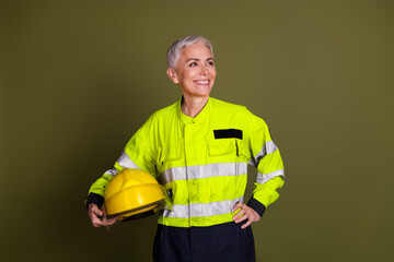 Mature woman in high-visibility workwear smiling confidently against a khaki backdrop, holding a yellow safety helmet