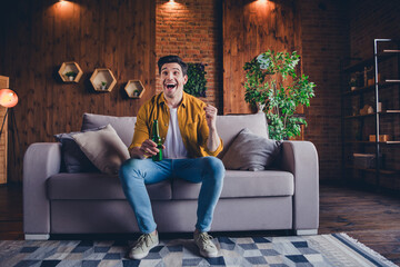 Excited young man relaxing indoors on a weekend, holding a drink and smiling in his comfortable homey loft living room with stylish decor