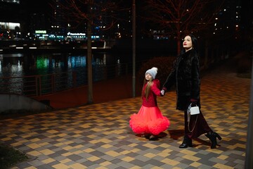 Woman in black coat walks hand in hand with girl in red dress along lit path by river at night