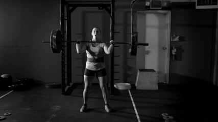 Dramatic black and white shot of young female power lifter lifting barbell over her head in grungy gym.