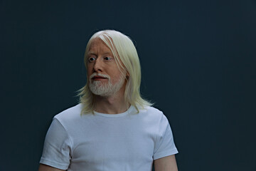 Portrait of a man with long white hair and a beard, wearing a plain white t shirt against a dark...